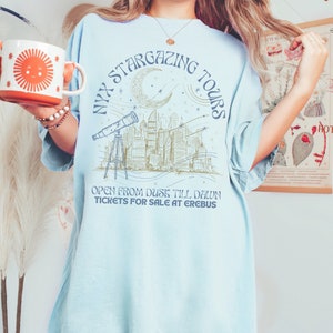K&ouml;nnte beinhalten: Ein hellblaues T-Shirt mit einem grafischen Design einer Skyline, einem Halbmond, Sternen und einem Teleskop. Der Text "NYX STARGAZING TOURS" ist in einem Kreis um das Design geschrieben. Der Text "OPEN FROM DUSK TILL DAWN TICKETS FOR SALE AT EREBUS" ist unter dem Design geschrieben.