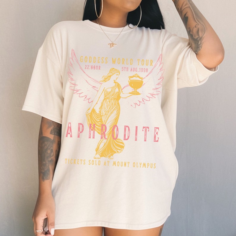 May include: A white t-shirt with a graphic print of a woman with wings holding a cup. The text on the shirt reads "Goddess World Tour 22/56HR 5TH AUG.1998 Aphrodite Tickets Sold at Mount Olympus".