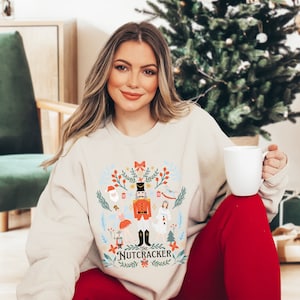 May include: A beige sweatshirt with a festive design featuring a Nutcracker, a ballerina, and other Christmas characters. The text "The Nutcracker" is printed on the bottom of the design.