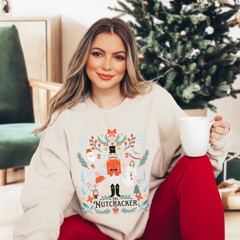 May include: A beige sweatshirt with a festive design featuring a Nutcracker, a ballerina, and other Christmas characters. The text "The Nutcracker" is printed on the bottom of the design.