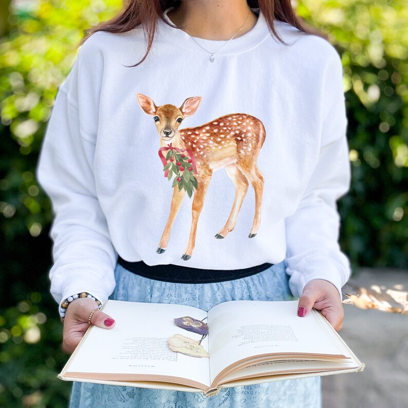 May include: White sweatshirt with a watercolor illustration of a fawn with a red bow and green leaves. The fawn is standing on a white background.
