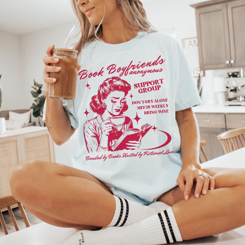 May include: A light blue t-shirt with a red graphic of a woman reading a book and crying. The text on the shirt reads "Book Boyfriends Anonymous Support Group Don't Cry Alone Meets Weekly Bring Wine Bonded by Books. United by Fictional Love."