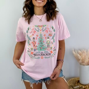 May include: Light pink t-shirt featuring a festive design from "The Nutcracker." The graphic includes a Christmas tree, nutcrackers, and floral elements. The text "The Nutcracker" is centered below the tree. A perfect holiday themed tee.