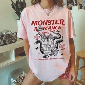 Pu&ograve; includere: T-shirt rosa chiaro con la scritta "MONSTER ROMANCE BOOK CLUB" in rosso. La grafica presenta una pila di libri, tentacoli, cuori e la frase "MATED. MARKED. MENTALLY UNWELL."