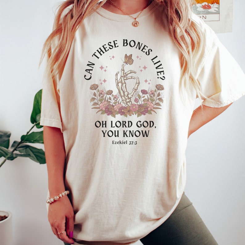 Puede incluir: Una camiseta beige con un estampado gr&aacute;fico de una mano esquel&eacute;tica sosteniendo una mariposa. El texto dice "Can these bones live? Oh Lord God, you know. Ezekiel 37:3".