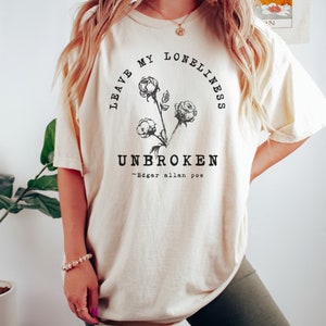 May include: A cream-colored t-shirt with a black graphic design featuring a floral motif and the text "Leave My Loneliness Unbroken - Edgar Allan Poe".