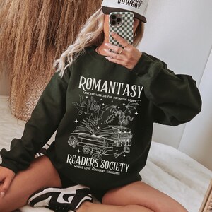 May include: Black sweatshirt with a white graphic design that says "Romantasy: Fantasy Worlds for Romantic Souls" and "Readers Society: Where Love Conquers Kingdoms". The graphic features a winged cherub, a dragon, and a stack of books.