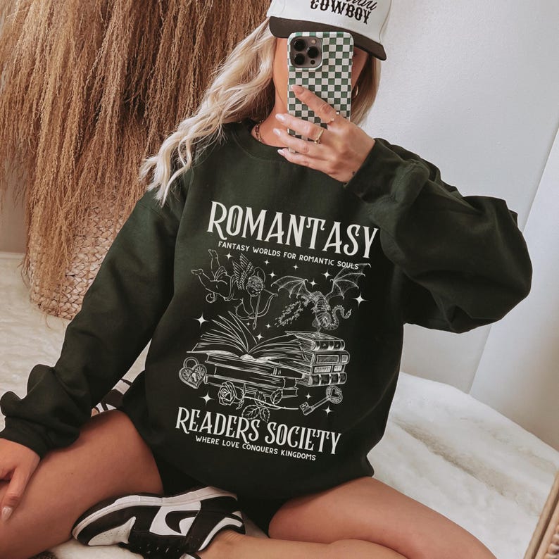 May include: Black sweatshirt with a white graphic design that says "Romantasy: Fantasy Worlds for Romantic Souls" and "Readers Society: Where Love Conquers Kingdoms". The graphic features a winged cherub, a dragon, and a stack of books.