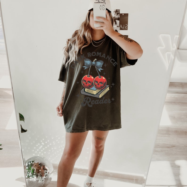 K&ouml;nnte beinhalten: Dunkel olivgr&uuml;nes T-Shirt mit einem Aufdruck von zwei roten Kirschen mit Totenkopfgesichtern, einer blauen Schleife und dem Text "Dark Romance Reader". Das Design ist auf einem weichen, &uuml;bergro&szlig;en T-Shirt gedruckt.