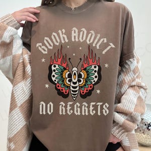 May include: Brown t-shirt with the text "BOOK ADDICT NO REGRETS" and a butterfly design. The butterfly has black, teal, red, and yellow wings with flames. The shirt is a crew neck and has short sleeves.