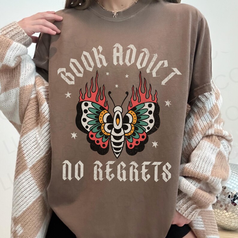 May include: Brown t-shirt with the text "BOOK ADDICT NO REGRETS" and a butterfly design. The butterfly has black, teal, red, and yellow wings with flames. The shirt is a crew neck and has short sleeves.
