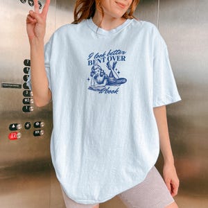 K&ouml;nnte beinhalten: Hellblaues T-Shirt mit dem marineblauen Text "I look better bent over a book". Die Grafik zeigt eine Vintage-Illustration einer lesenden Frau. Das Shirt hat eine lockere Passform.