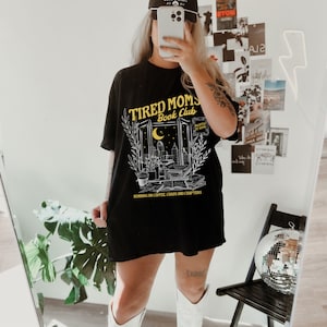 May include: Black t-shirt featuring a graphic of a window with a cityscape and the text "Tired Moms Book Club" in yellow. The text below the graphic reads "Running on coffee, chaos and chapters."