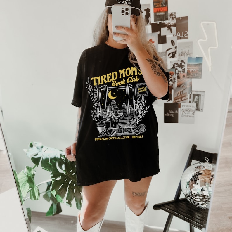 May include: Black t-shirt featuring a graphic of a window with a cityscape and the text "Tired Moms Book Club" in yellow. The text below the graphic reads "Running on coffee, chaos and chapters."