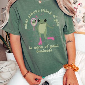 May include: Green t-shirt with a frog wearing pink boots and looking in a mirror. The text on the shirt reads "What others think of you is none of your business."