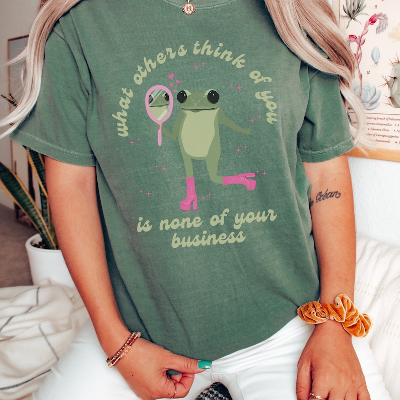 May include: Green t-shirt with a frog wearing pink boots and looking in a mirror. The text on the shirt reads "What others think of you is none of your business."