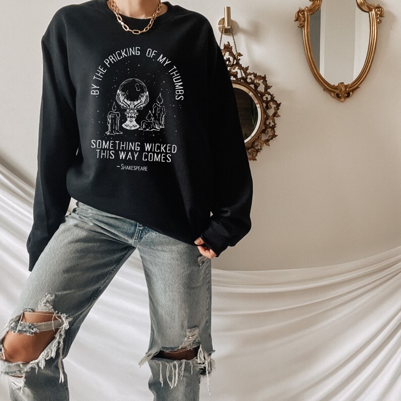 Op de afbeelding: Zwart sweatshirt met een witte grafische print van een kristallen bol, kaarsen en de tekst "By the pricking of my thumbs, something wicked this way comes - Shakespeare".