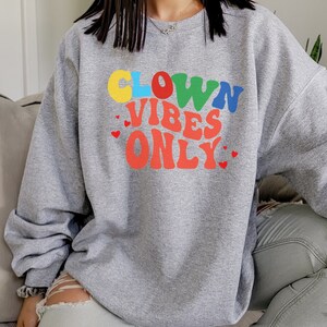 Może przedstawiać: Szara bluza z kolorowym nadrukiem, na kt&oacute;rym widnieje napis "Clown Vibes Only" w retro czcionce. Nadruk zawiera r&oacute;wnież małe czerwone serduszka.