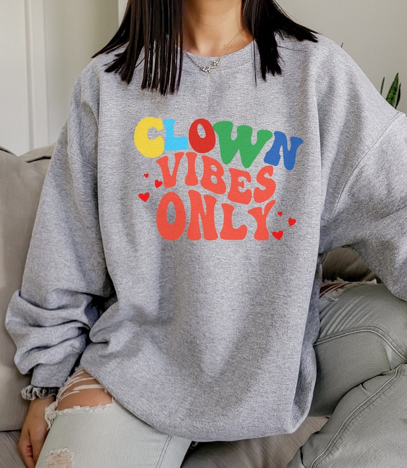 Może przedstawiać: Szara bluza z kolorowym nadrukiem, na kt&oacute;rym widnieje napis "Clown Vibes Only" w retro czcionce. Nadruk zawiera r&oacute;wnież małe czerwone serduszka.