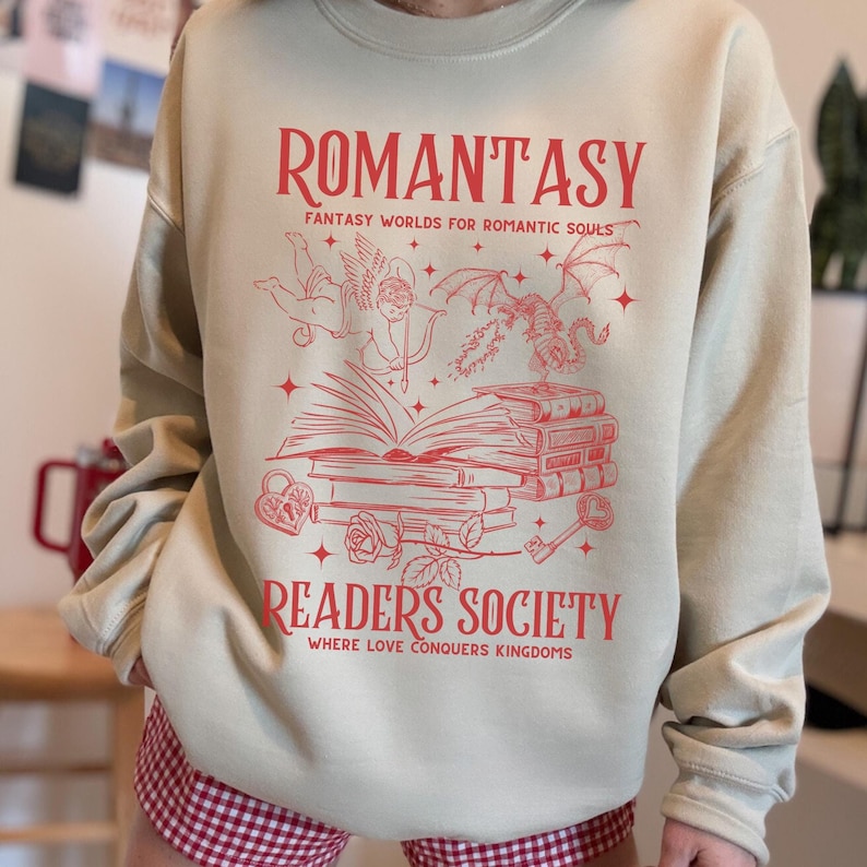 May include: A beige sweatshirt with a red graphic design that says "Romantasy: Fantasy Worlds for Romantic Souls" and "Readers Society: Where Love Conquers Kingdoms". The graphic features a winged cupid, a dragon, a book, and a key.