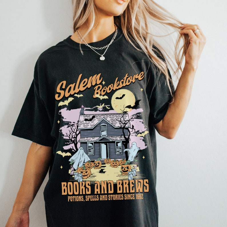 Op de afbeelding: Zwart T-shirt met een afbeelding van een spookhuis, vleermuizen en geesten. De tekst "Salem Bookstore" staat boven het huis en "Books and Brews" staat onder het huis. De tekst "Potions, Spells and Stories Since 1992" staat onder de tekst "Books and Brews".