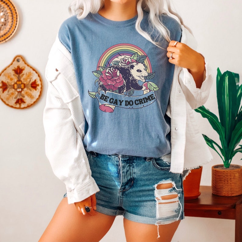 May include: A blue t-shirt featuring a possum, roses, and a rainbow design. The text "BE GAY DO CRIME" is displayed on a banner. The shirt is paired with denim shorts.