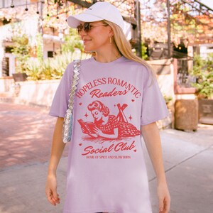 Peut inclure: Un t-shirt lavande avec une illustration vintage d'une femme lisant un livre. Le texte sur le t-shirt est "Hopeless Romantic Readers Social Club, Home of Spice and Slow Burn".