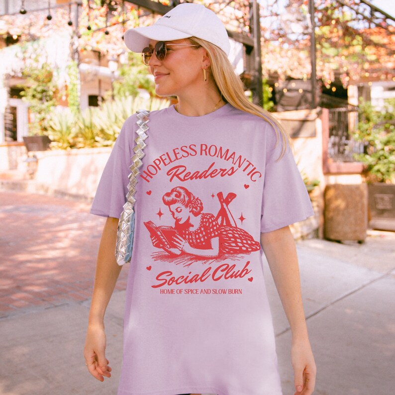 Peut inclure: Un t-shirt lavande avec une illustration vintage d'une femme lisant un livre. Le texte sur le t-shirt est "Hopeless Romantic Readers Social Club, Home of Spice and Slow Burn".