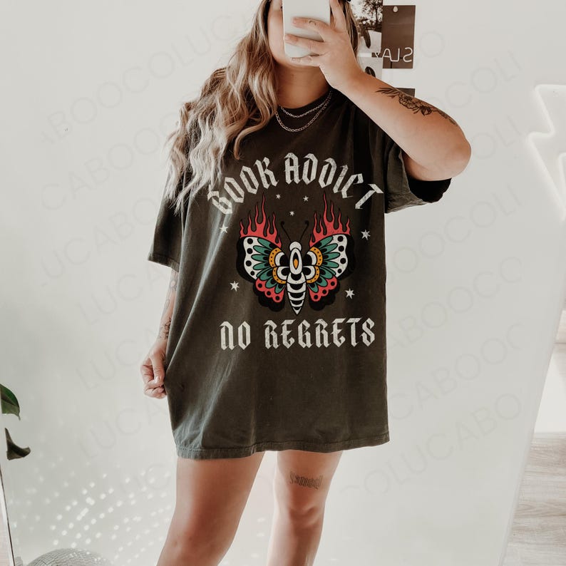 May include: Dark green t-shirt with the words "BOOK ADDICT" above a butterfly design with red, yellow, and teal accents. Below the butterfly is the phrase "NO REGRETS" in white lettering. The shirt is a casual style.