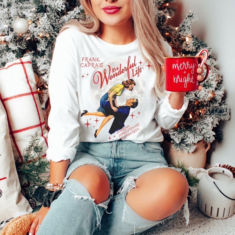Puede incluir: Camiseta blanca de manga larga con un gr&aacute;fico de una pareja bailando de la pel&iacute;cula "&iexcl;Qu&eacute; bello es vivir!" El texto "Frank Capra's It's a Wonderful Life" est&aacute; impreso en la camiseta. La camiseta la lleva puesta una persona que sostiene una taza roja con el texto "merry + bright".