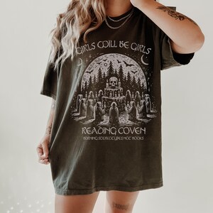 Może przedstawiać: Ciemnozielona koszulka z napisem "GIRLS WILL BE GIRLS" na g&oacute;rze. Poniżej grafika przedstawiająca czaszkę, drzewa i postacie, z napisami "READING COVEN" i "BURNING STEREOTYPES, HOT BOOKS".