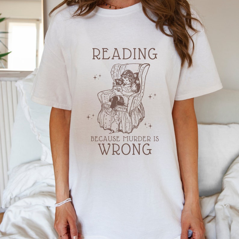 Puede incluir: Camiseta blanca con una ilustraci&oacute;n marr&oacute;n de una ni&ntilde;a leyendo en una silla. El texto "READING BECAUSE MURDER IS WRONG" est&aacute; impreso debajo de la ilustraci&oacute;n.