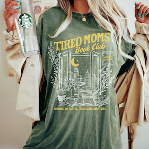 May include: Green t-shirt with a white graphic design of a window with a cityscape, books, and a crescent moon. The text "Tired Moms Book Club" is in yellow above the window and "Nightly at 10" is in yellow to the right of the window. The text "Running on coffee, chaos and chapters" is below the window.