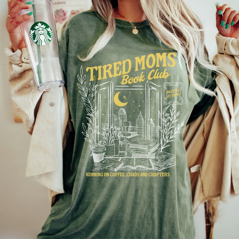 May include: Green t-shirt with a white graphic design of a window with a cityscape, books, and a crescent moon. The text "Tired Moms Book Club" is in yellow above the window and "Nightly at 10" is in yellow to the right of the window. The text "Running on coffee, chaos and chapters" is below the window.
