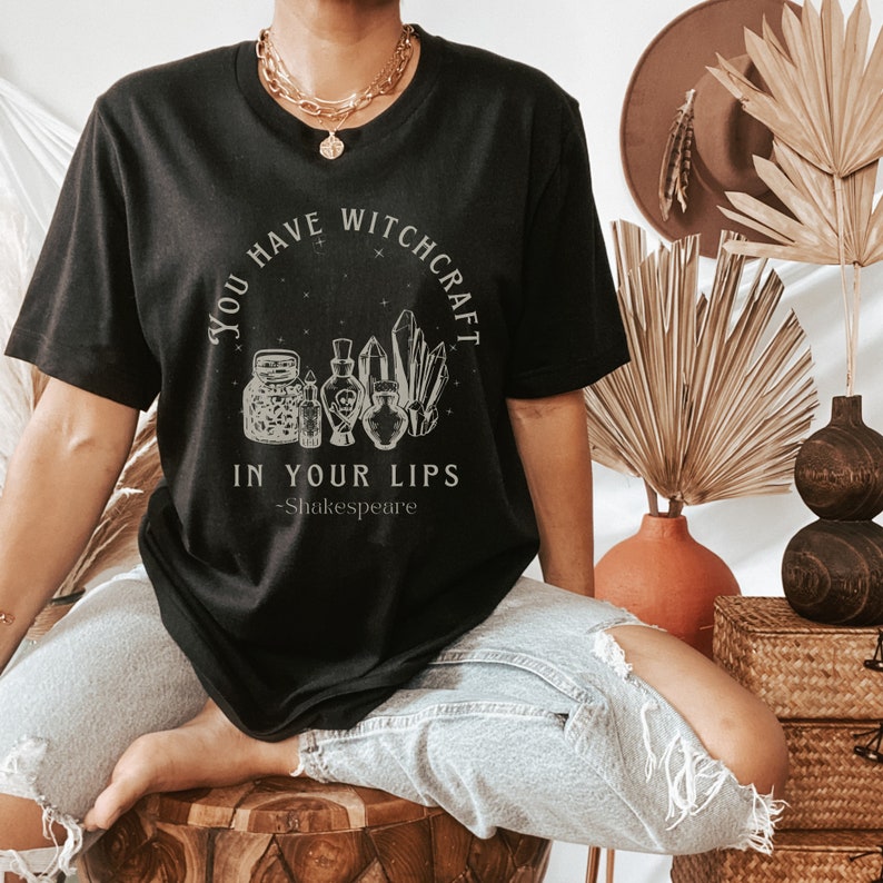Op de afbeelding: Zwart T-shirt met een witte grafische print van flessen en kristallen met de tekst "You have witchcraft in your lips -Shakespeare".