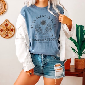 Op de afbeelding: Een blauw T-shirt met een grafisch ontwerp met een zon, palmbladeren en de tekst "To be great is to be misunderstood - Ralph Waldo Emerson".