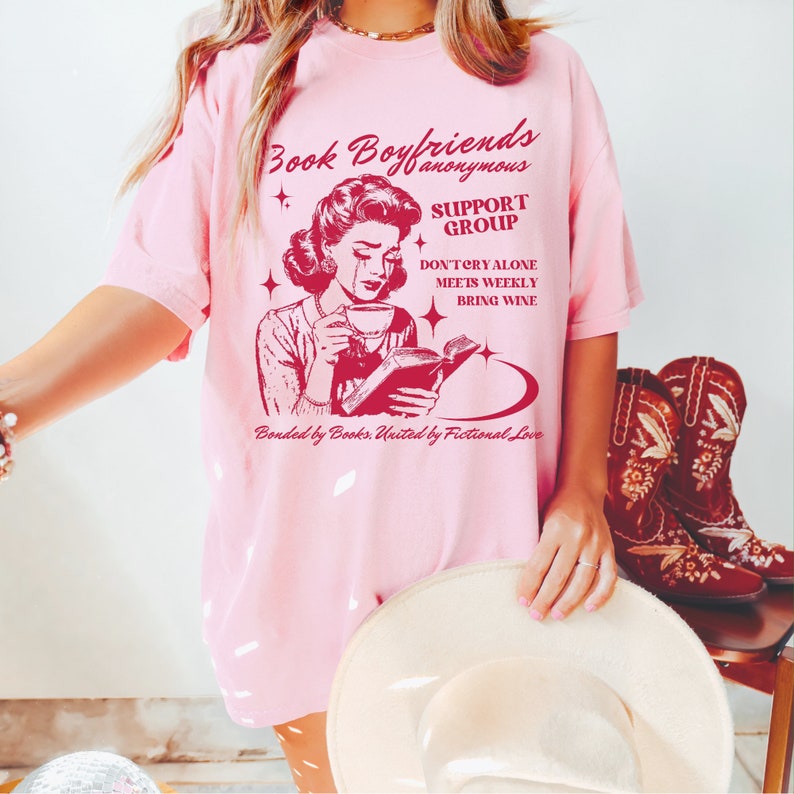 May include: Pink t-shirt with a red graphic of a woman reading a book and crying. The text on the shirt reads "Book Boyfriends Anonymous Support Group Don't Cry Alone Meets Weekly Bring Wine Bonded by Books. United by Fictional Love."