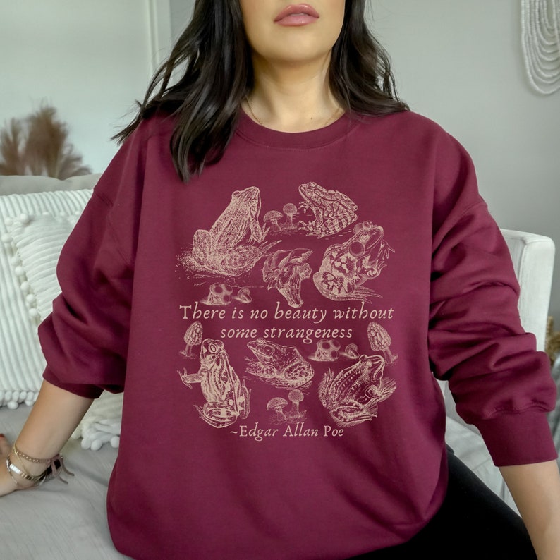 May include: Burgundy sweatshirt with a graphic print of frogs and mushrooms. The text "There is no beauty without some strangeness - Edgar Allan Poe" is printed on the sweatshirt.