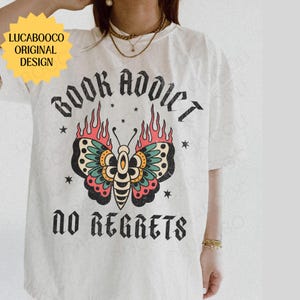 May include: Cream-colored t-shirt featuring a colorful butterfly design with flames and the words "BOOK ADDICT" and "NO REGRETS." The shirt also has the text "LUCABOOCO ORIGINAL DESIGN" in a yellow starburst.