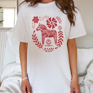 May include: White t-shirt featuring a red Scandinavian Dala horse design. The horse is surrounded by red floral patterns.
