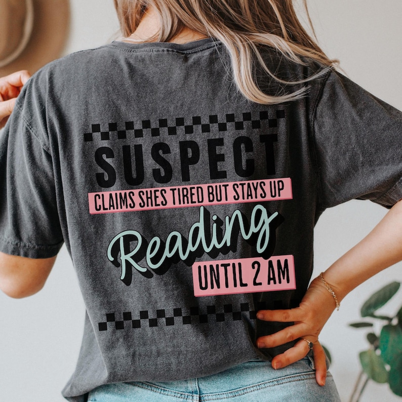 May include: A black t-shirt with a white and black checkered pattern at the top and bottom. The text "SUSPECT" is in black, "CLAIMS SHES TIRED BUT STAYS UP" is in pink, "Reading" is in light blue, and "UNTIL 2 AM" is in pink.