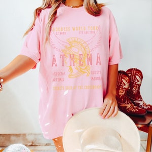Puede incluir: Camiseta rosa de gran tama&ntilde;o con un gr&aacute;fico de una cabeza de mujer con un casco con alas. El texto en la camiseta dice "GODDESS WORLD TOUR", "ATHENA", "22:06NR", "5TH AUG.1982", "+SPECIAL ARTEMIS", "GUESTS MEDUSA", y "TICKETS SOLD AT THE CROSSROADS".