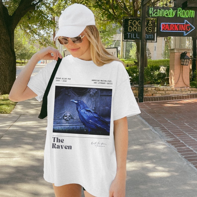 May include: White t-shirt with a graphic print of a raven on a tombstone. The text "The Raven" is printed below the image. The text "Edgar Allan Poe 1809 - 1849" and "American Writer, Poet, and Literary Critic" is printed above the image.