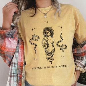 May include: A yellow t-shirt with a black and white graphic of a woman with snakes and the text "STRENGTH BEAUTY POWER".