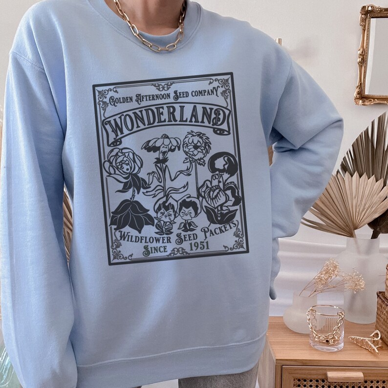 May include: Light blue sweatshirt with a black graphic print of a vintage seed packet design. The text on the packet reads "Golden Afternoon Seed Company Wonderland Wildflower Seed Packets Since 1951".
