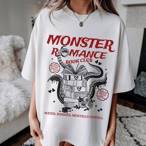 Pu&ograve; includere: T-shirt color crema con la scritta rossa "MONSTER ROMANCE BOOK CLUB". Il design presenta una pila di libri, tentacoli e cuori. &Egrave; presente anche il testo "Mated. Marked. Mentally Unwell."