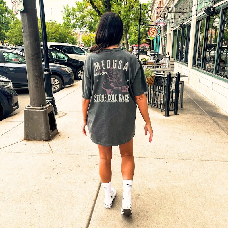 May include: A person wearing a dark gray t-shirt with a graphic print of Medusa and the text "Medusa and the Gorgons World Tour Stone Cold Gaze".