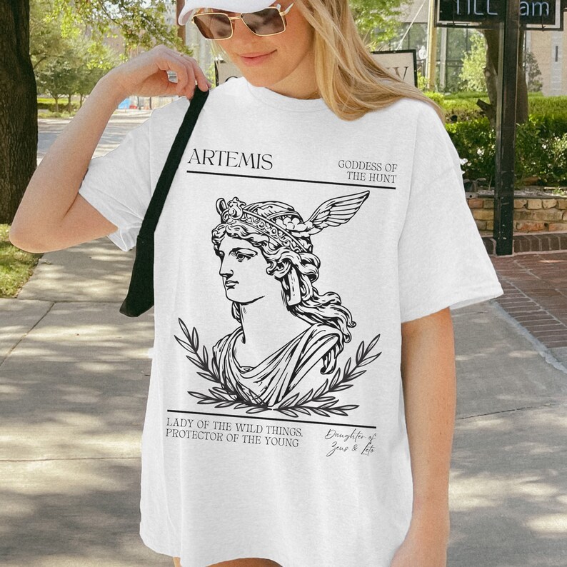 May include: White t-shirt with a black and white illustration of Artemis, the Greek goddess of the hunt. The text reads "Artemis, Goddess of the Hunt, Lady of the Wild Things, Protector of the Young, Daughter of Zeus & Leto."