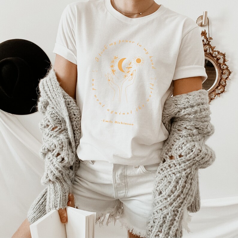 May include: White t-shirt with a yellow graphic design featuring a hand holding a crescent moon and sun with a quote from Emily Dickinson: "I took my power in my hand and went against the world."