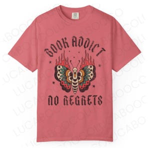 May include: A dusty rose t-shirt with the words "BOOK ADDICT" above a butterfly design with flames and the words "NO REGRETS" below. The butterfly has red, green, and yellow details.